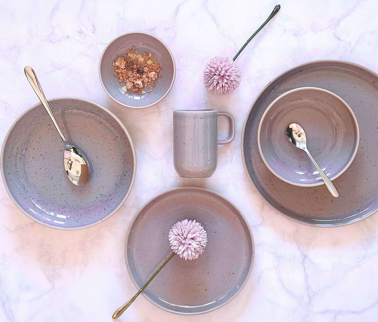 Ein hellgraues, gesprenkeltes Mäser Tafelservice »Candy« mit Tellern, Schalen und Tasse auf Marmor. Eine Schale enthält Müsli, zwei Löffel und zwei rosa Blumen sind ebenfalls zu sehen.