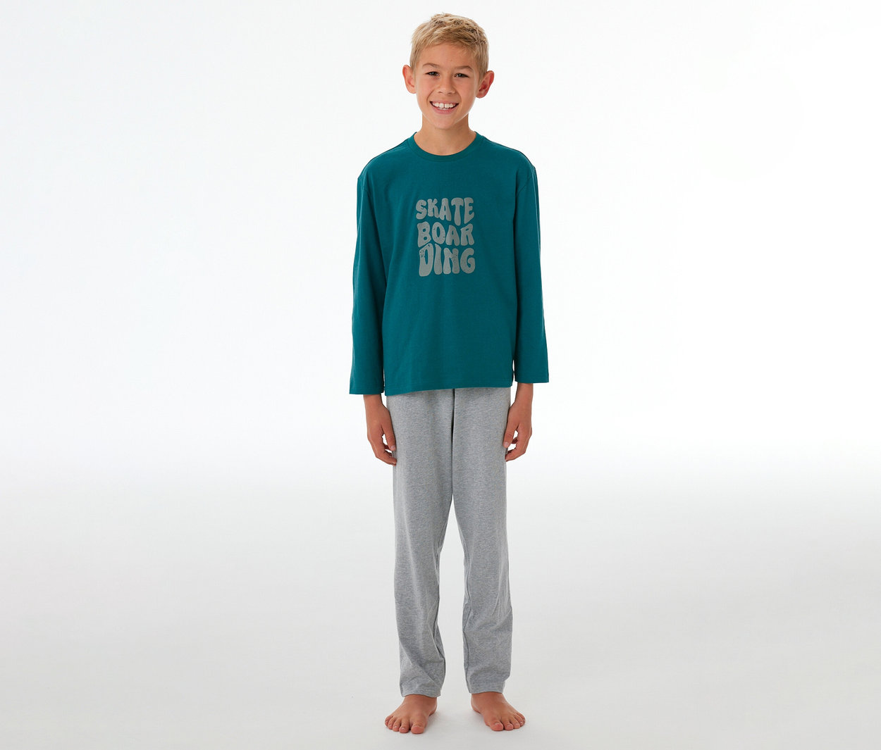 Lächelnder Junge in einem Kinder-Pyjama mit petrolfarbenem Langarmshirt, grauer "SKATE BOARDING"-Aufschrift und grauer Pyjamahose.