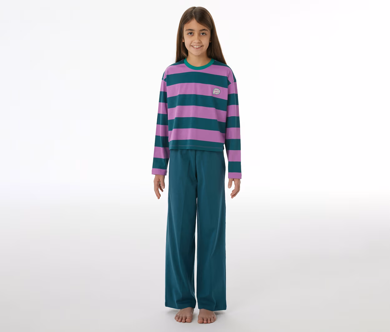 Mädchen lächelt, trägt einen SCHIESSER Kinder-Pyjama mit lila-grün gestreiftem Langarmshirt, grüner Hose und hat nackte Füße.