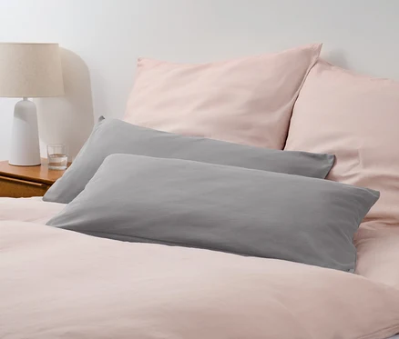 Zwei graue Jersey-Kopfkissenbezüge liegen auf einem Bett mit rosa Bettwäsche.