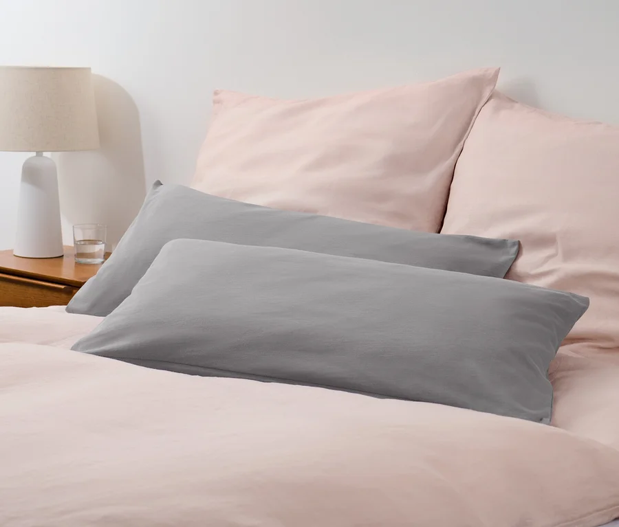 Zwei graue Jersey-Kopfkissenbezüge liegen auf einem Bett mit rosa Bettwäsche.