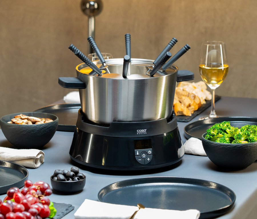 Tisch mit Caso Induktions-Fondue, Tellern, Schüsseln mit Essen, Weinglas und Trauben.