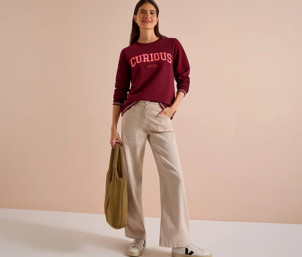 Lächelnde Frau in einem bordeauxroten CECIL Sweatshirt mit „CURIOUS“-Aufschrift, beiger Hose und weißen Sneakern, die eine olivgrüne Tasche hält.