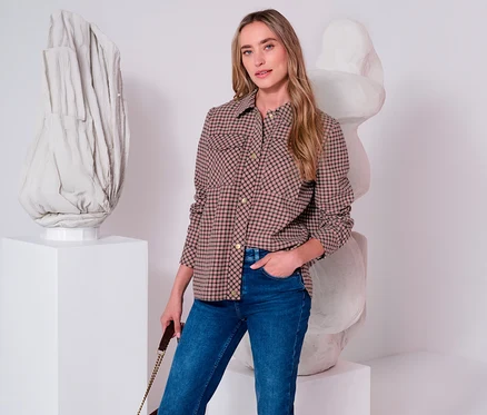 Eine Frau trägt eine braun-schwarz karierte Street One Jacke und blaue Jeans, mit der Hand in der Tasche, vor abstrakten Skulpturen.