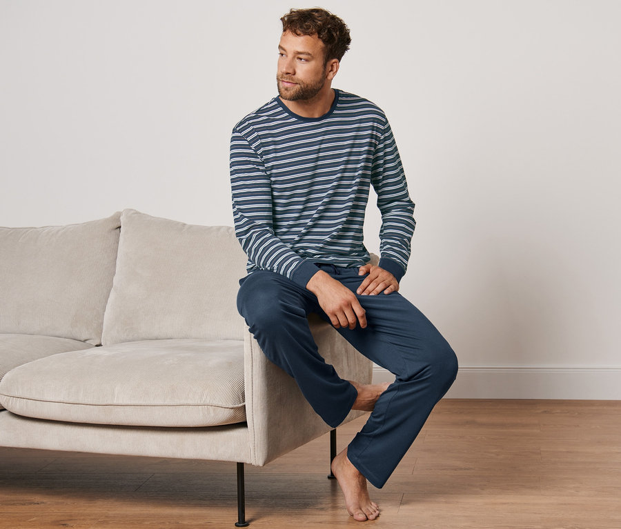 Mann sitzt auf beigem Sofa, trägt blauen, gestreiften Pyjama mit Modal.