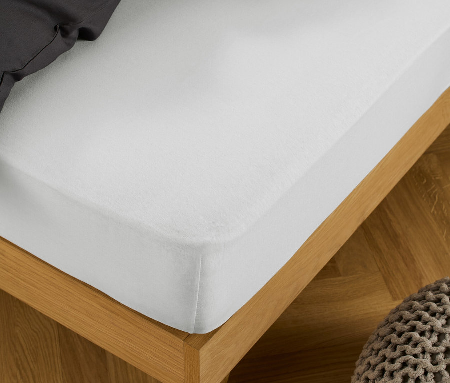 Ein weißes Jersey-Spannbettlaken ist auf ein Bett mit Holzrahmen gespannt, daneben steht ein Strickhocker.