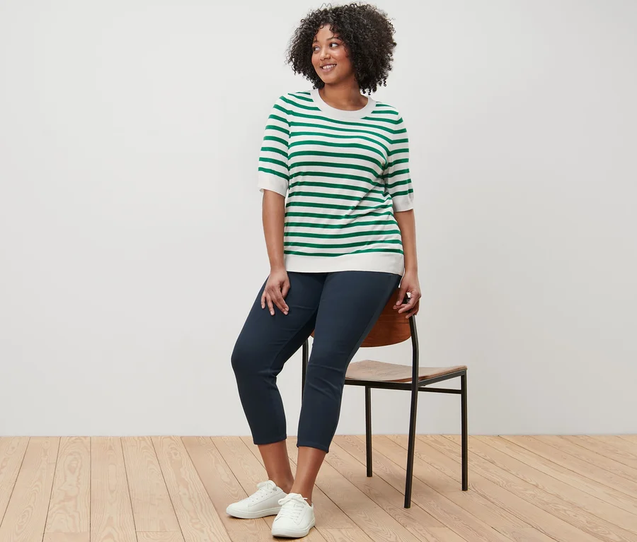 Frau sitzt auf einem Stuhl und trägt einen gestreiften Feinstrickpullover und eine 7/8-Jeggings.