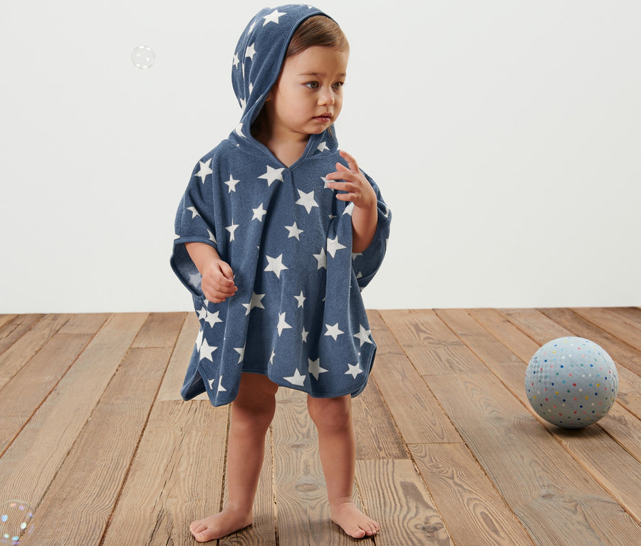 Ein Kind steht auf einem Holzboden und trägt einen blauen Frottierponcho mit Kapuze und weißen Sternen.