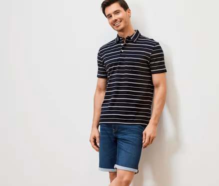 Mann lächelt an eine Wand gelehnt. Er trägt ein dunkelblau gestreiftes Jersey-Poloshirt und Jeansshorts.