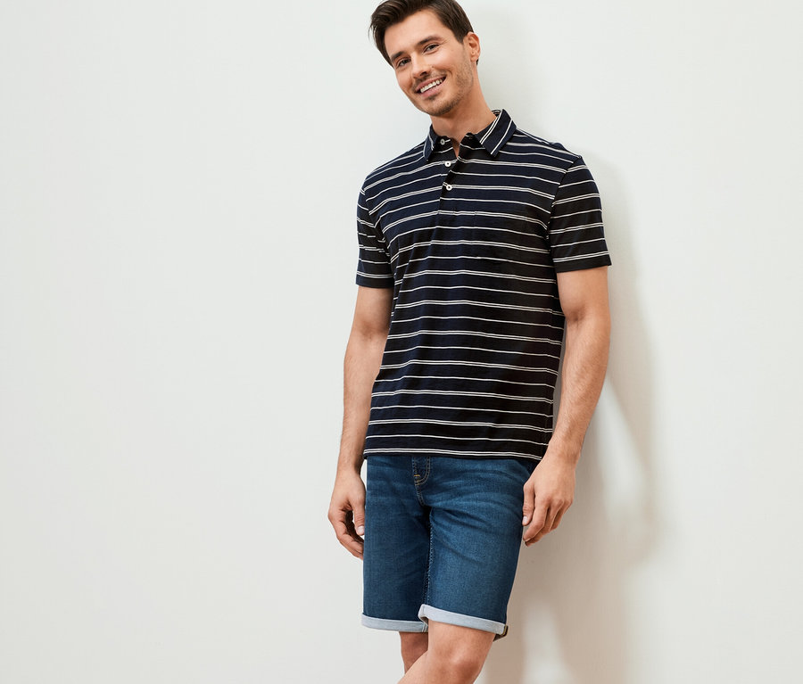 Mann lächelt an eine Wand gelehnt. Er trägt ein dunkelblau gestreiftes Jersey-Poloshirt und Jeansshorts.