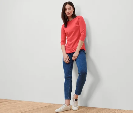 Frau posiert in einem korallenfarbenen Shirt mit 3/4-Arm, blauen Jeans und weißen Turnschuhen.