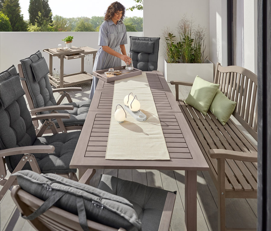 Eine Frau gießt Wasser auf einer Terrasse mit einem Leira Ausziehtisch, Hochlehnern, LED-Akku-Tischleuchten und einer Leira Bank mit hochklappbarem Tabletttisch.