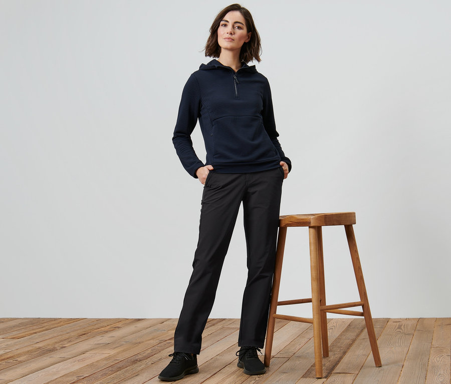 Frau posiert neben einem Holzhocker und trägt eine Softshellhose.