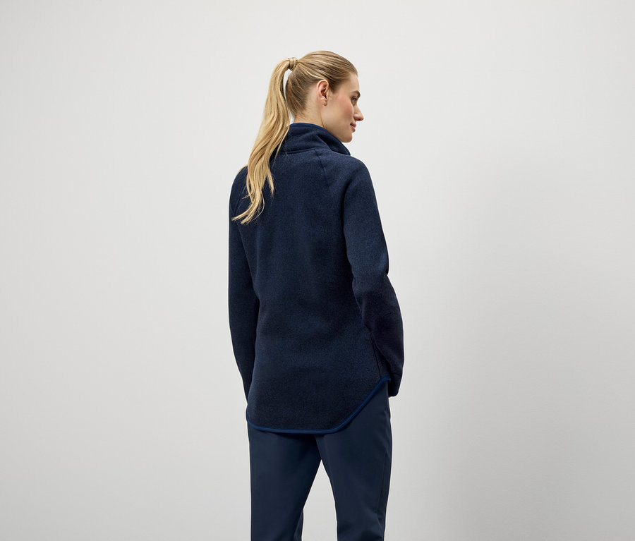 Von hinten ist eine Frau mit blonden, zu einem Pferdeschwanz zusammengebundenen Haaren zu sehen, die einen dunkelblauen Pullover und eine Hose trägt.