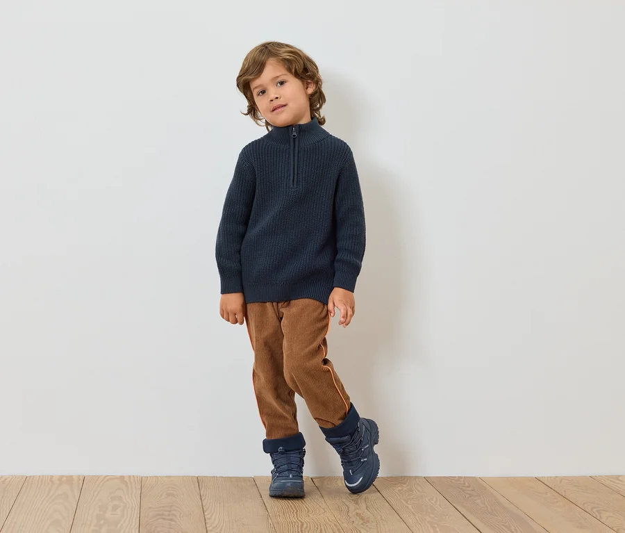 Ein Junge steht vor einer weißen Wand. Er trägt einen dunkelblauen Pullover, braune Hosen und blaue Schuhe.