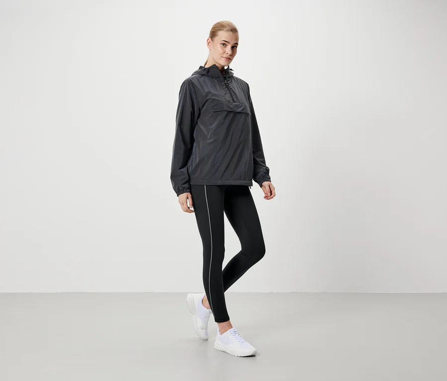 Blonde Frau steht in grauer Kapuzenjacke, schwarzen Leggings und weißen Schuhen.