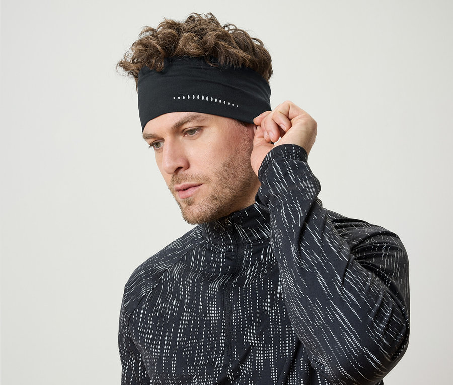 Mann mit lockigem Haar justiert ein schwarzes Sportstirnband mit weißen Details. Er trägt ein schwarzes Langarmshirt.