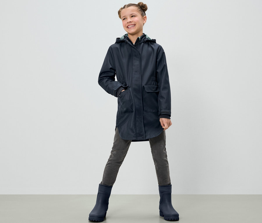 Ein lächelndes Mädchen posiert in einem Kinder-Softshellmantel, grauen Hosen und blauen Stiefeln.