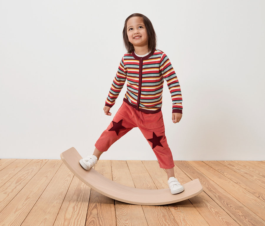 Kind steht auf einem hölzernen Balance Board, trägt einen gestreiften Pullover und Hosen mit Sternen.