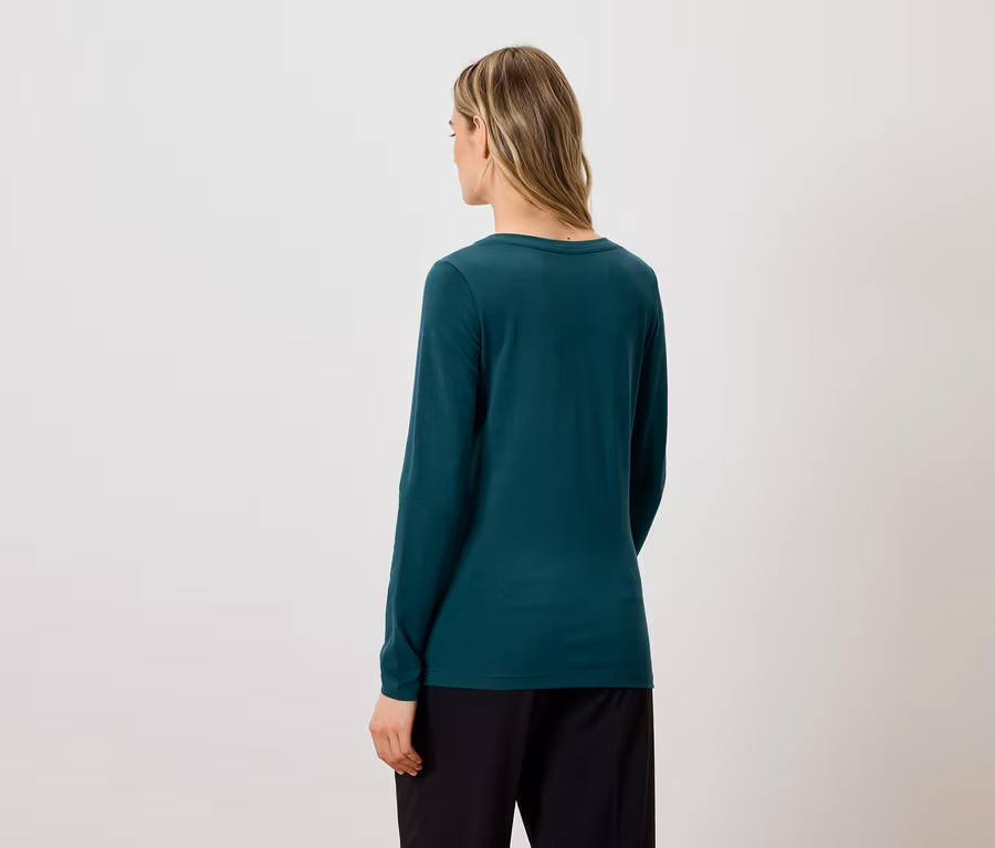 Von hinten ist eine Frau mit blonden Haaren zu sehen, die ein grünes Langarmshirt und eine schwarze Hose trägt.