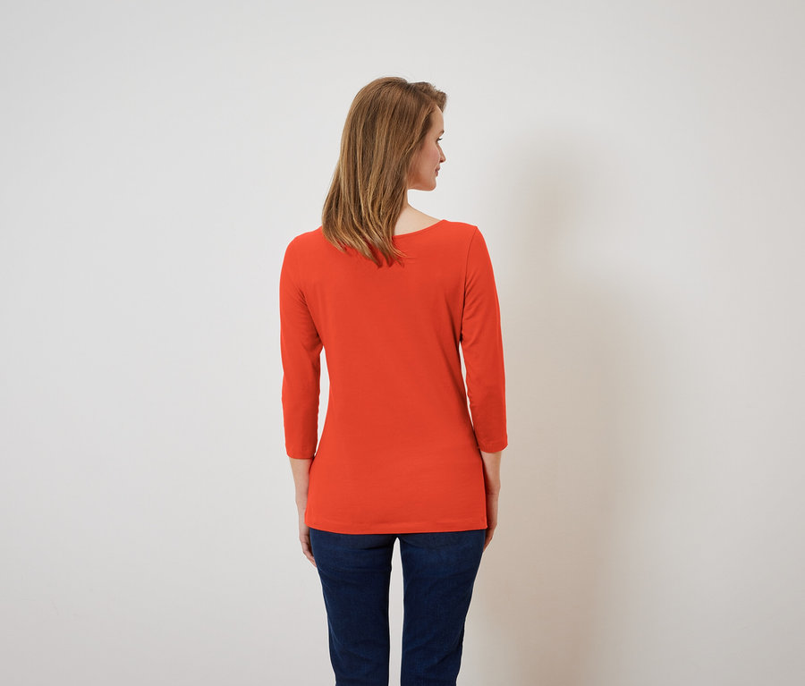 Frau mit dem Rücken zur Kamera trägt ein orangefarbenes Dreiviertelarmshirt und dunkelblaue Jeans.