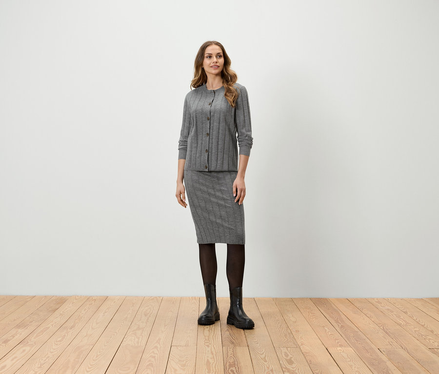 Eine Frau steht und trägt eine graue Strickjacke, einen Rock, eine schwarze Strumpfhose und Stiefel.