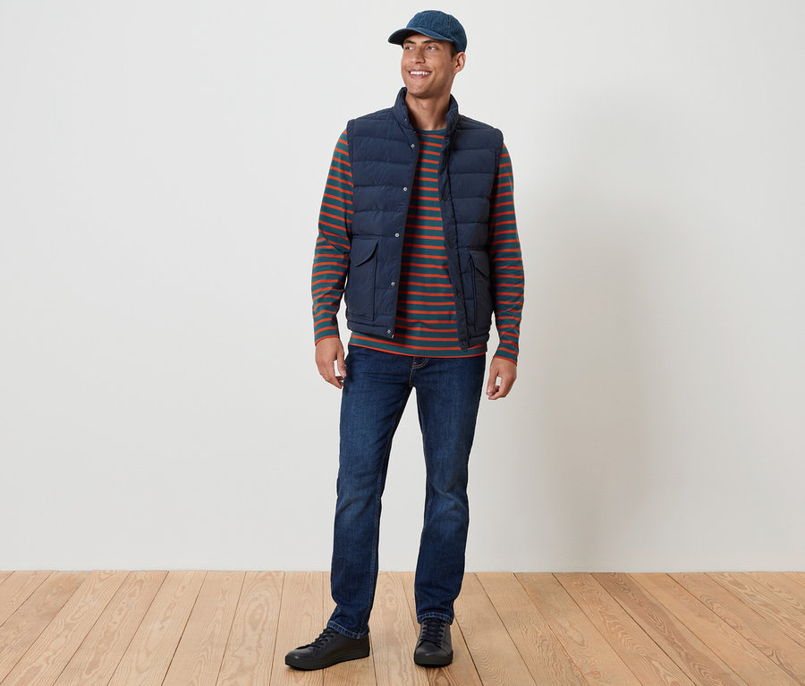 Ein Mann steht und lächelt. Er trägt eine blaue Weste, ein gestreiftes T-Shirt, Jeans und eine blaue Kappe.