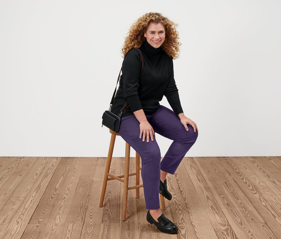 Frau sitzt auf einem Hocker und trägt einen schwarzen Feinstrick-Rollkragenpullover, lila Hosen und schwarze Schuhe.