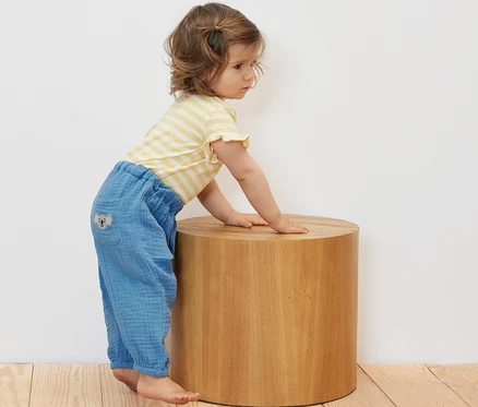 Kleinkind lehnt in Baby-Musselin-Schlupfhose an einem Holzhocker.