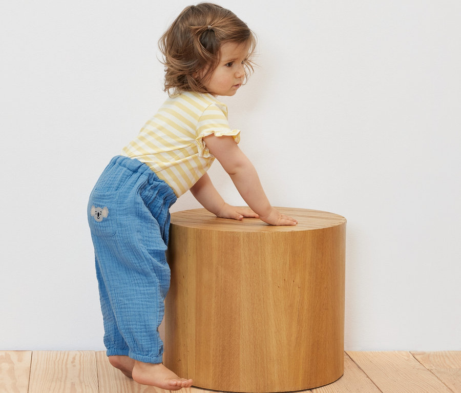 Kleinkind lehnt in Baby-Musselin-Schlupfhose an einem Holzhocker.