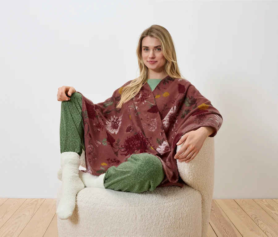 Eine blonde Frau sitzt in einem Sessel und trägt einen geblümten Kimono, eine grüne Hose und weiße Socken.