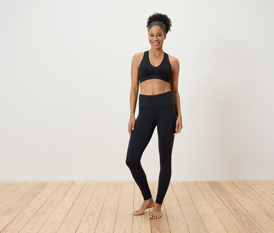 Eine Frau in einem schwarzen Sport-BH und Leggings steht auf einem Holzboden vor einer weißen Wand.
