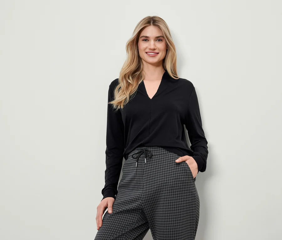 Blonde Frau in schwarzem Langarmshirt und grauer Hahnentrittmusterhose posiert.