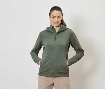 Frau mit olivgrünem Kapuzenpullover mit Reißverschluss und beigen Hosen, die mit beiden Händen in den Taschen steht.