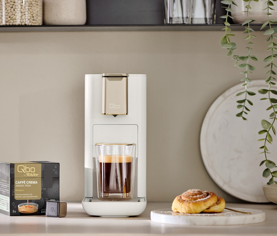 Eine Qbo ICONIC Light Gold Kaffeemaschine mit einer Tasse Kaffee daneben. Eine Packung Qbo Kapseln und ein Zimtgebäck sind auch zu sehen.