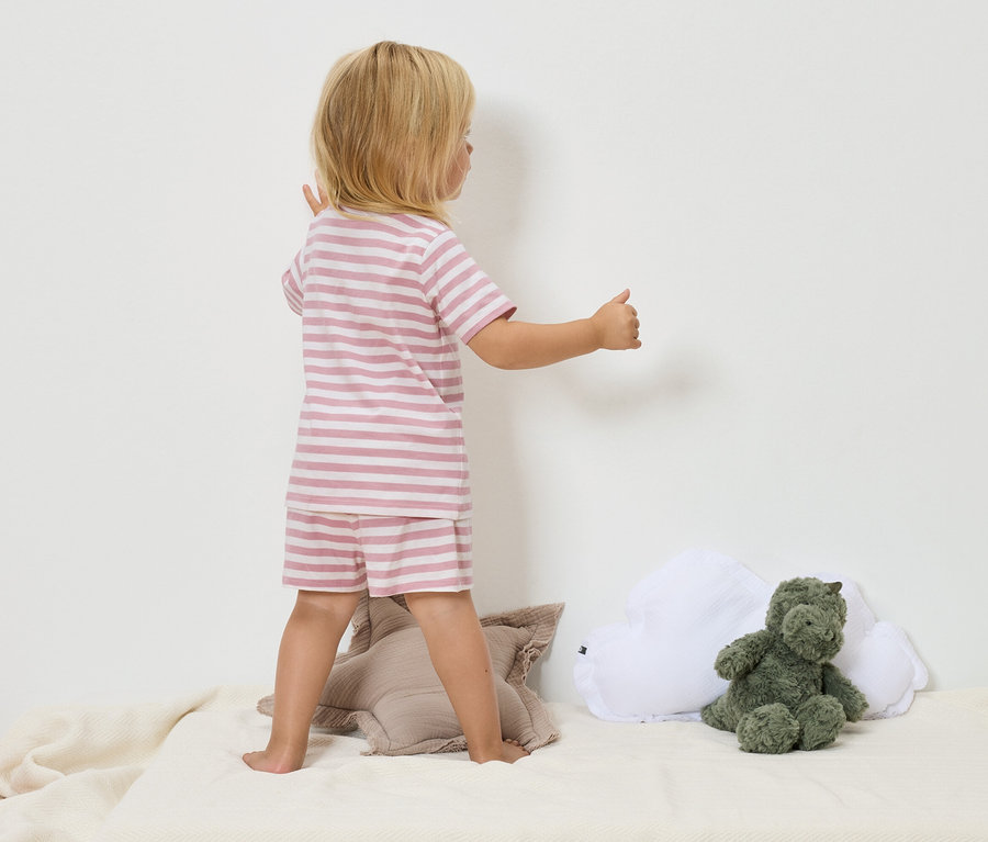 Ein Kind mit blonden Haaren, das einen gestreiften rosa-weißen Pyjama trägt, steht auf einer beigen Decke neben einem grünen Dinosaurier-Stofftier und einem weißen Wolkenkissen.