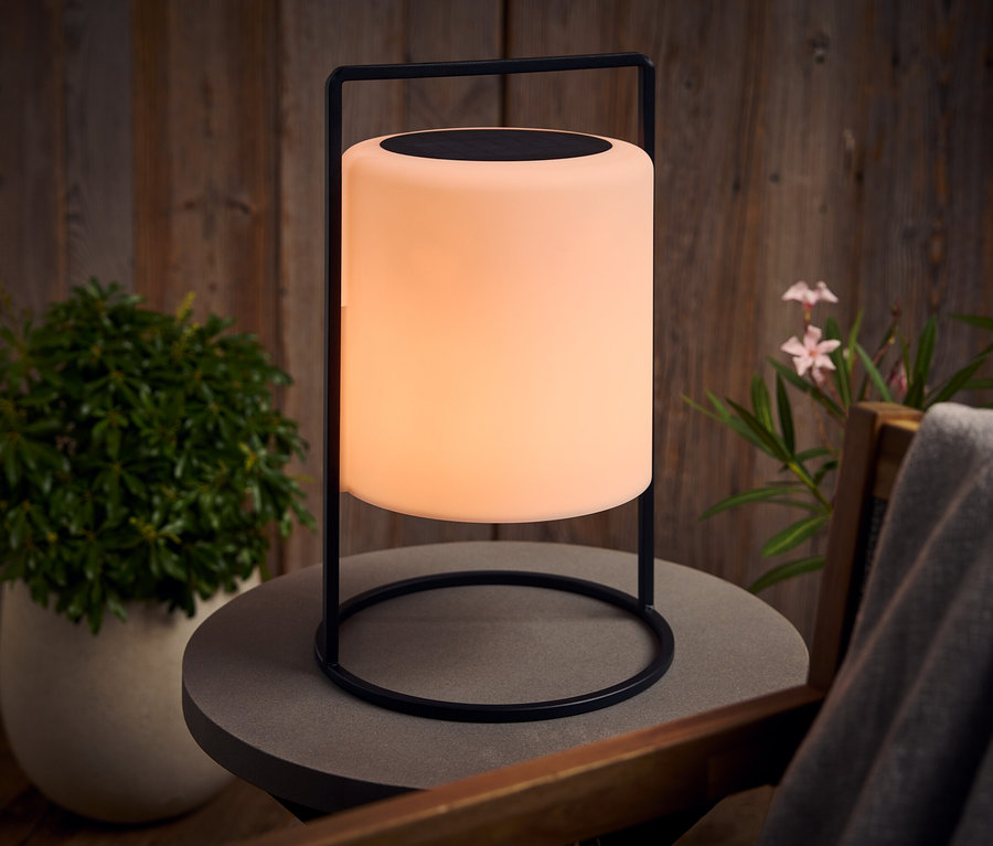 Eine leuchtende Lampe mit schwarzem Gestell steht auf einem grauen Tisch im Freien.