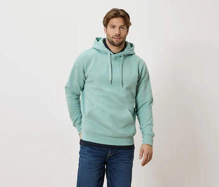 Mann mit helltürkisem Hoodie und Jeans steht vor einem weißen Hintergrund.
