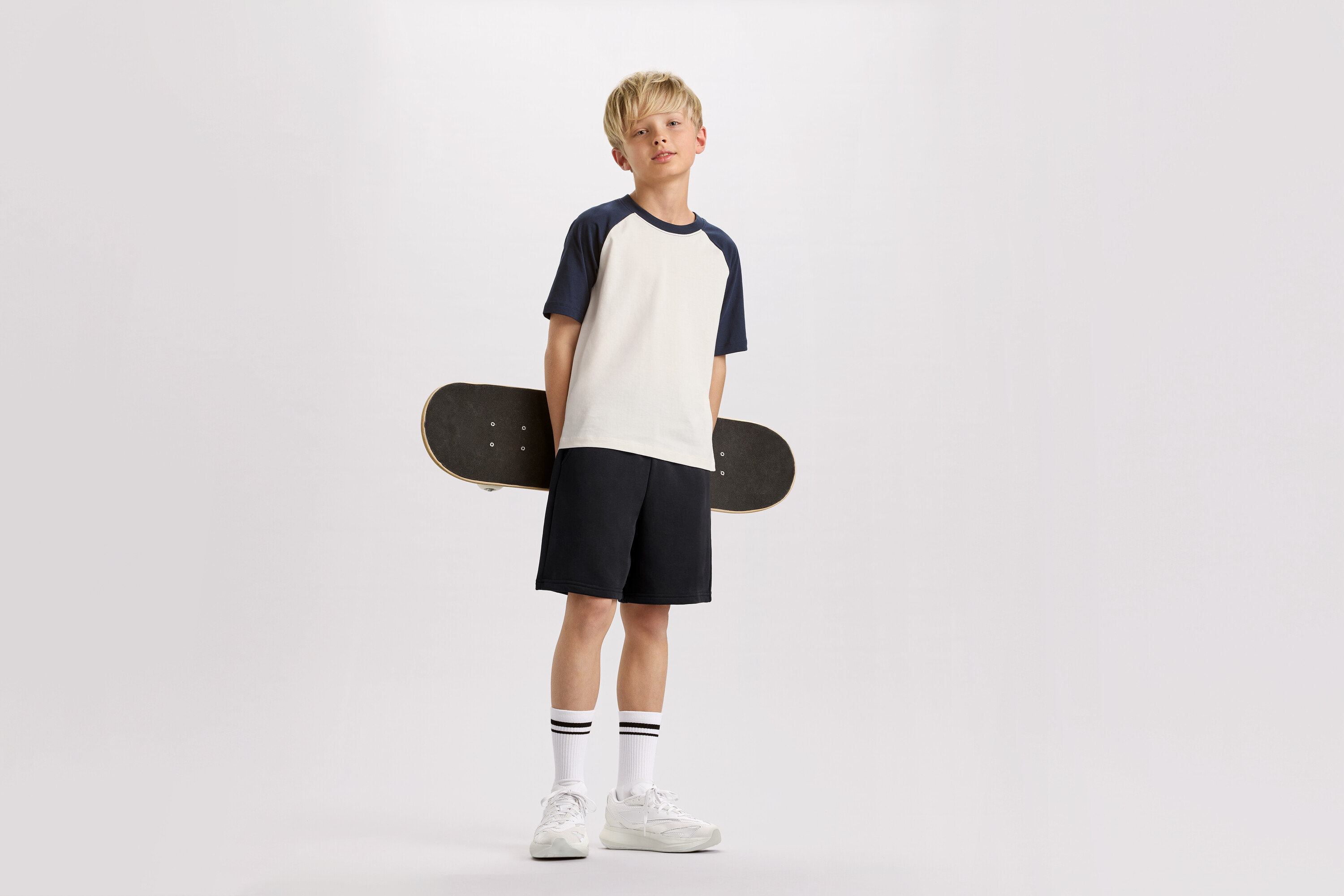 Ein blonder Junge hält ein Skateboard hinter seinem Rücken, trägt ein T-Shirt mit marineblauen Ärmeln, Shorts und Turnschuhe.