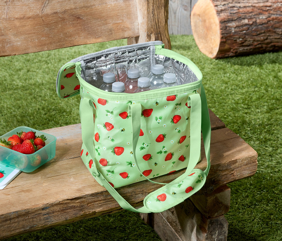 Eine hellgrüne Kühltasche mit Erdbeermuster steht auf einer Holzbank, daneben eine Schüssel mit Erdbeeren und Wasserflaschen in der Tasche.