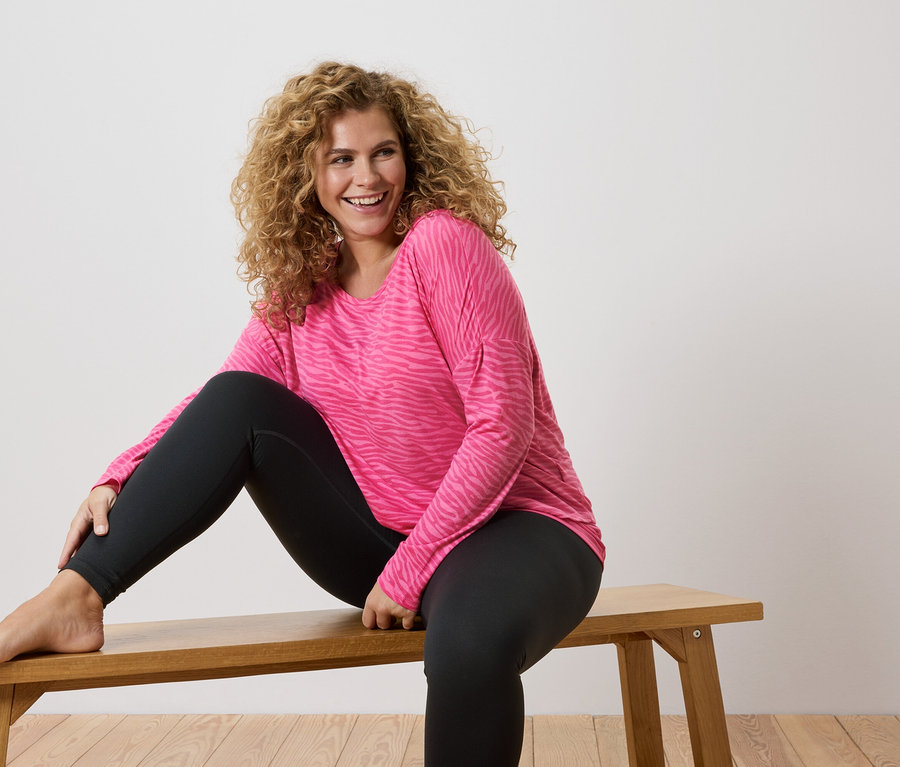 Frau mit lockigen Haaren sitzt barfuß auf einer Holzbank, trägt ein pinkes, langärmliges Shirt mit Zebramuster und schwarze Leggings.
