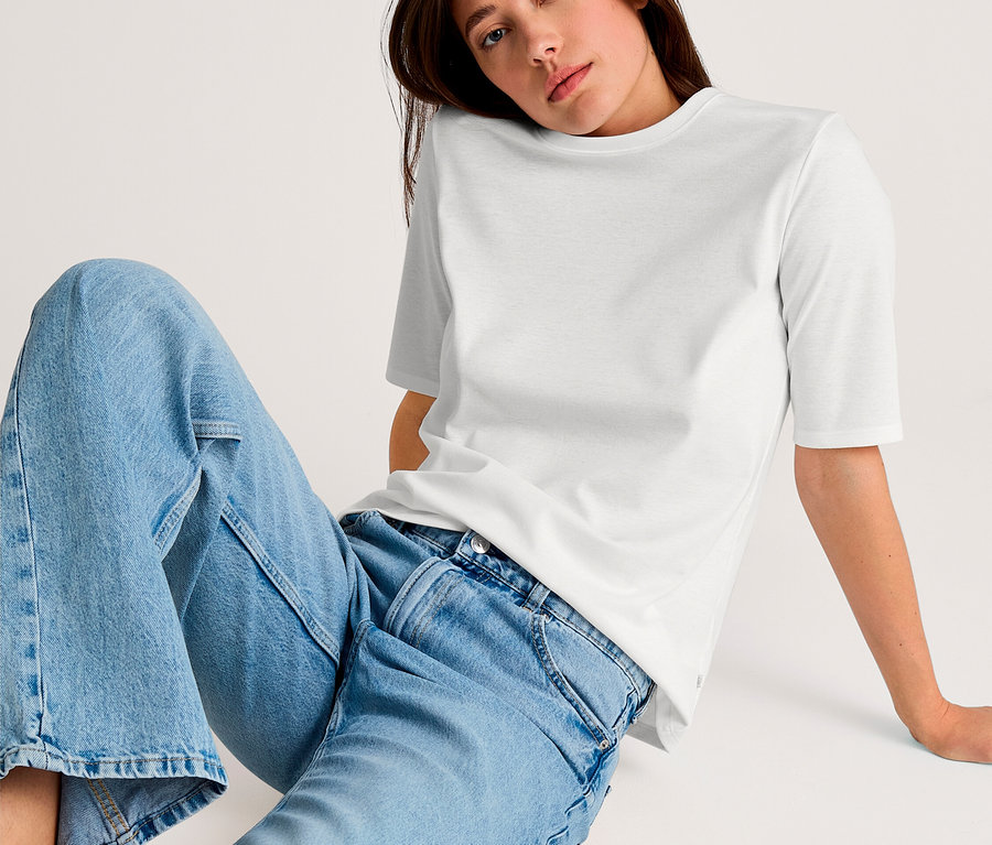Frau sitzt und trägt ein weißes T-Shirt und hellblaue Jeans.
