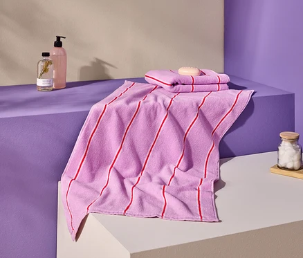 Aufnahme von rosa Handtüchern mit roten Streifen, Seife, Flaschen und Behältern auf einer lila und cremefarbenen Oberfläche.