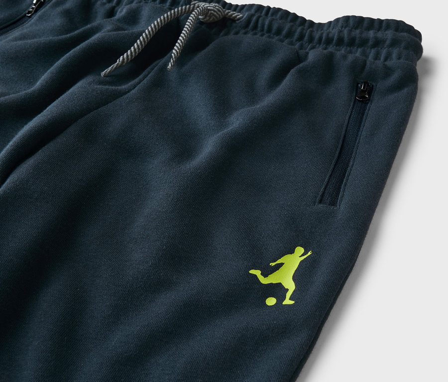 Nahaufnahme einer marineblauen Trainingshose mit Kordelzug, Reißverschlusstasche und hellgrünem Fußballspieler-Logo.