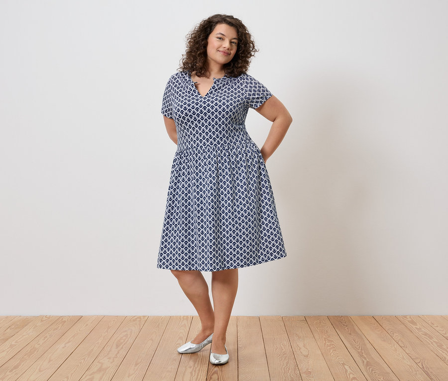 Frau mit lockigem Haar, die ein blau-weißes Kleid und silberne Schuhe trägt und mit den Händen in den Taschen vor einer Wand steht.