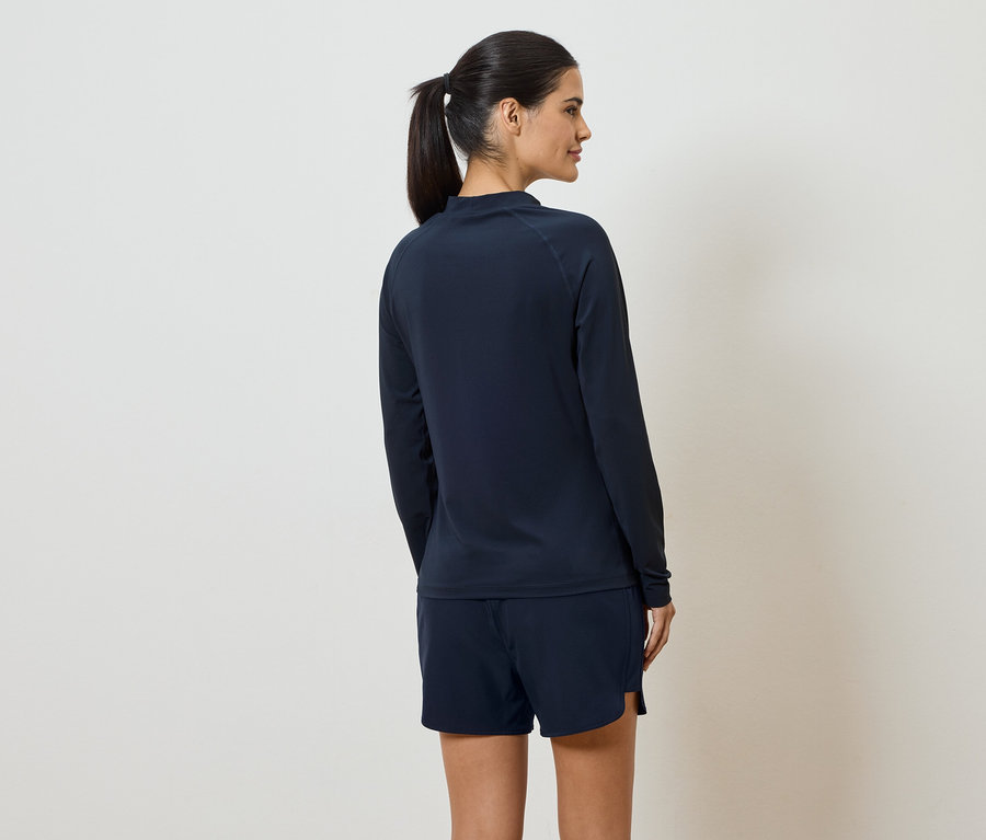 Eine Frau mit Pferdeschwanz trägt ein marineblaues Langarmshirt und Shorts, von hinten gesehen vor einem cremefarbenen Hintergrund.