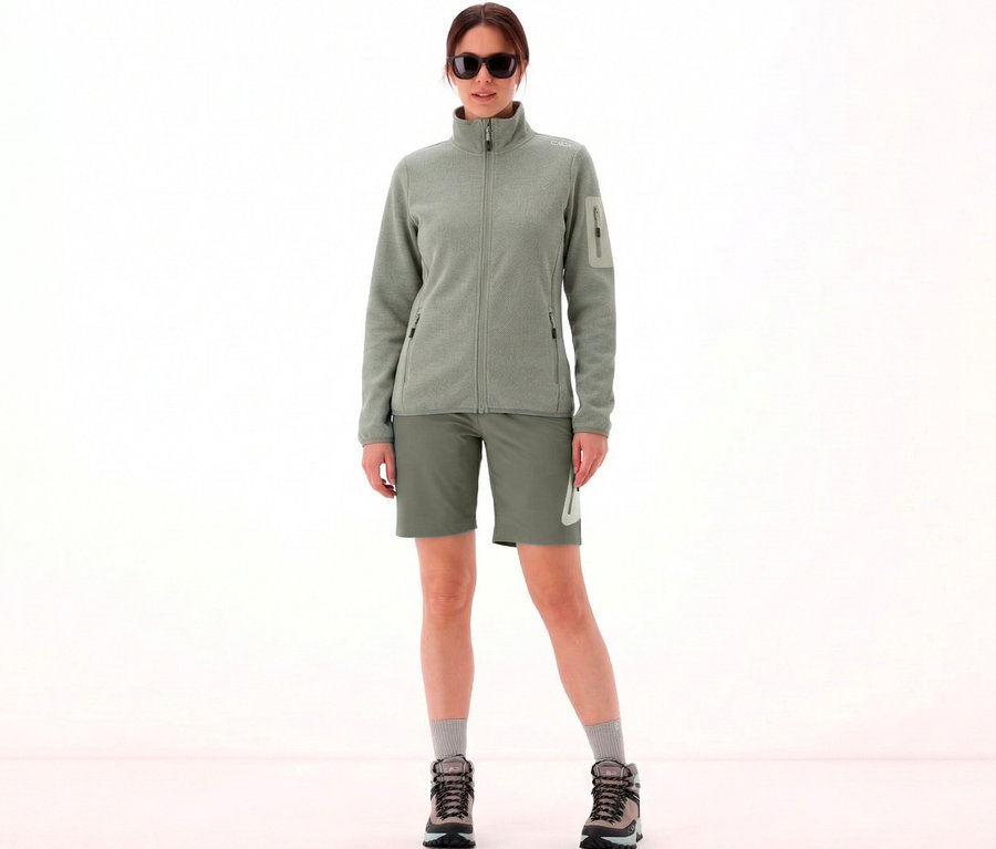 Frau mit Sonnenbrille trägt CMP Damen-Knit-Tech-Fleece in Avocado-Pistazie, Shorts und Wanderschuhe vor hellem Hintergrund.