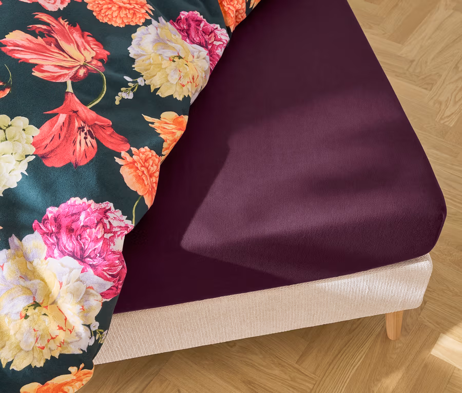 Nahaufnahme eines Bettes mit dunkelviolettem Spannbettlaken und einer Bettdecke mit Blumenmuster.