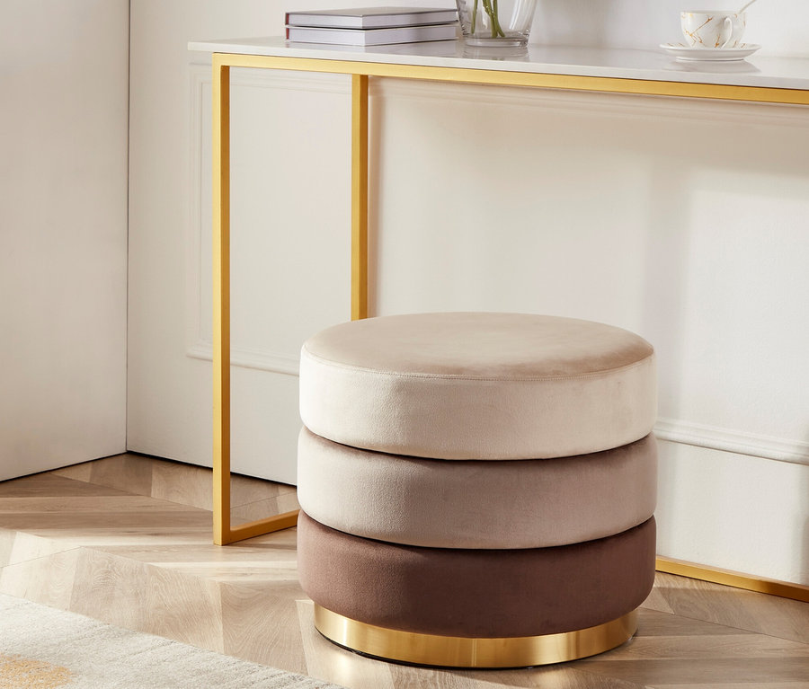 Ein dreifach gestapelter runder Hocker in Beige-, Braun- und Rosatönen steht neben einem goldenen Konsolentisch.