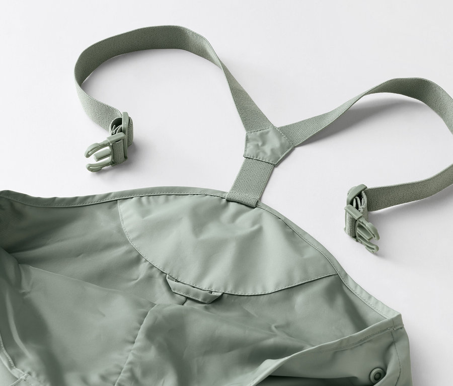 Nahaufnahme einer grünen Matschhose mit Trägern und Schnallen auf einem weißen Hintergrund.